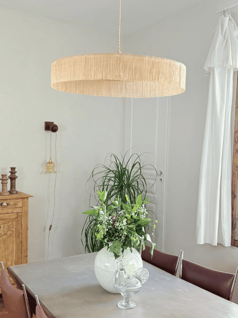 Grande suspension parasol à franges beiges vue de profil au dessus d'une table dans une décoration campagne chic. 